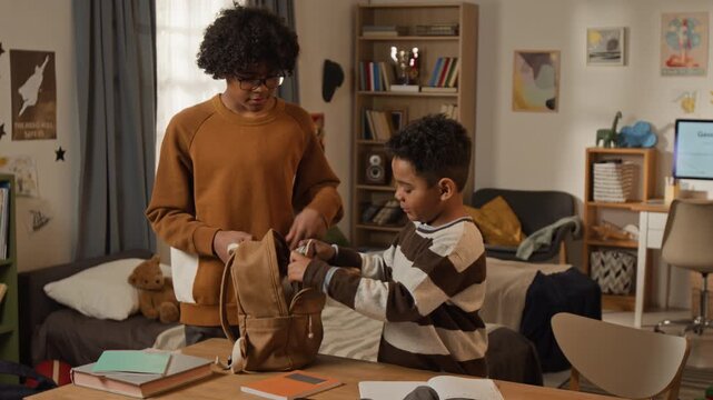 Medium shot of teenage African American boy helping little brother pack bag for first day at elementary school, holding while putting in textbooks, copybooks, stationery, then placing on shoulders