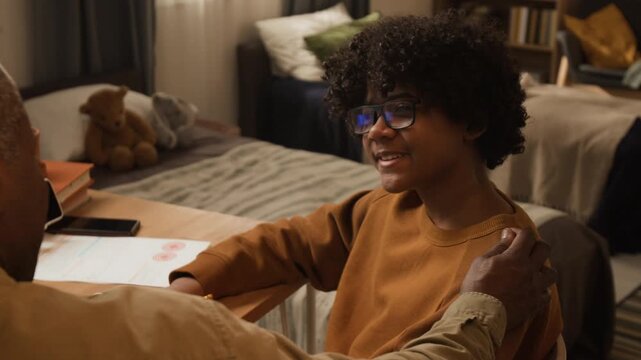 Medium close-up of proud teenage African American boy reporting study achievements and sharing good grades with supportive father, praising and patting son on shoulder
