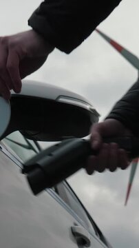 Vertical video of unrecognizable hand connects charging cable into EV car, wind charging station. Insert power connector to electric vehicle near sustainable electricity generator, wind turbine