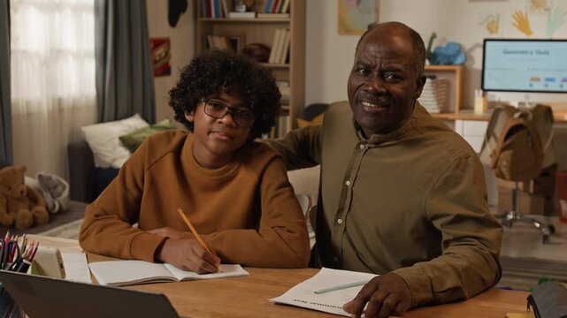 Medium portrait of cheerful mature African American father homeschooling teenage son, sitting at desk while boy is solving math in copybook, hugging, looking at camera, smiling, parental study support