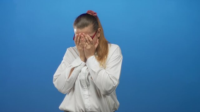 Woman in white shirt and red glasses covers face with both hands against a blue studio backdrop showing a gestural cry; distress vulnerability.