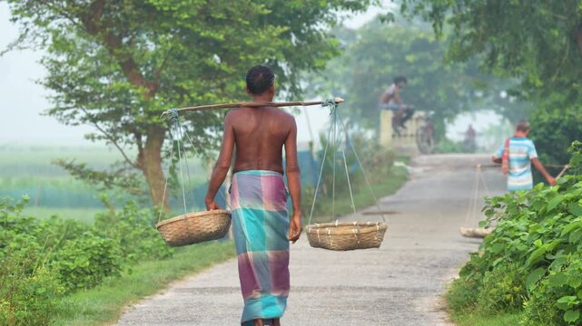 Bogura, Bangladesh - 22 September 2023: View of a man carrying baskets on a yoke while walking along a rural road surrounded by lush green trees and fields.