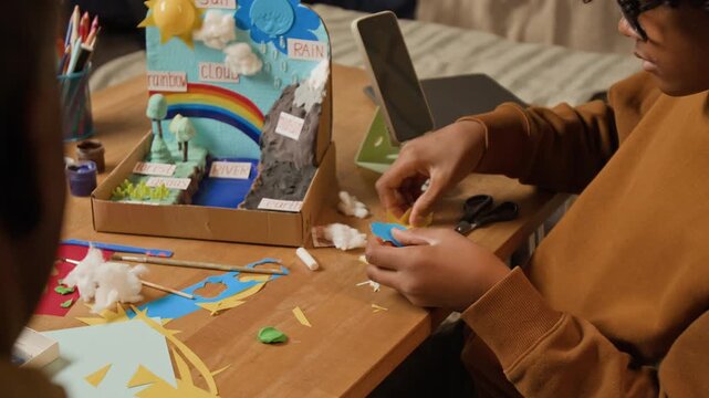 Over-shoulder shot of young black schoolboy making science project on earth climate, cutting out sun and cloud from color paper, while father is sitting by and guiding, representing child creativity