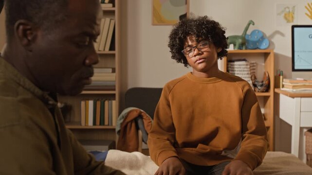Medium shot of strict black father packing schoolbag with lunchbox and snack for apathetic teenage son in bedroom in morning, then urging to hurry as boy refusing to go to school, for parent control