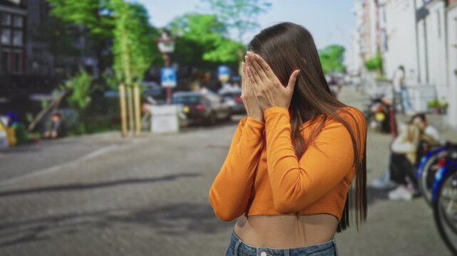 Young brunette woman covers face with both hands, exposed midriff and orange sweater visible on street near parked bikes and buildings; shyness solitude.