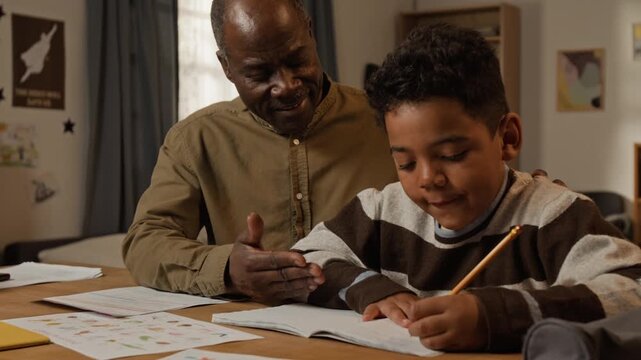 Medium shot of cheerful mature African American father helping young elementary age son with writing and spelling homework, smiling and encouraging while pointing to mistake in copybook