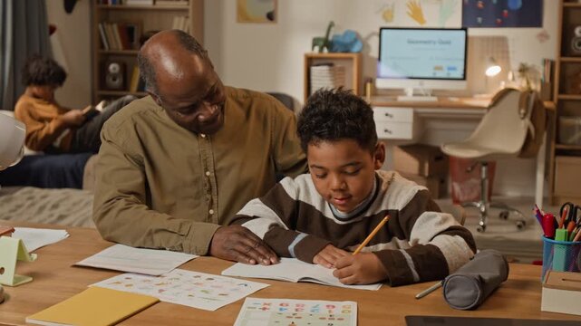 Medium shot of loving African American father and young son sitting together at desk, boy learning to write in copybook to prepare for elementary school while dad is stroking on head for encouragement