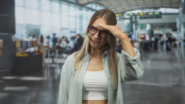 Woman wearing glasses shades eyes with hand in busy airport terminal, white crop top visible; travel waiting tired.
