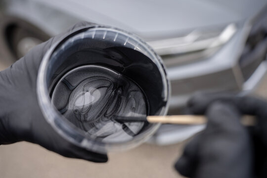 Hands in black nitrile gloves stirring silver metallic automotive paint in a plastic jar with a wooden stick, preparing car paint for refinishing or body repair service.