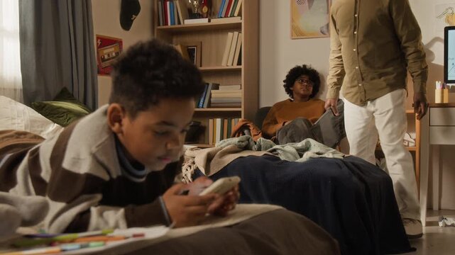 Medium full shot of black father taking away digital smartphone and tablet from lazy sons and requesting to clean up toys, children picking up construction blocks from floor, family discipline concept