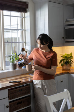 Stainless steel carafe pouring hot beverage into ceramic mug on wooden countertop near window