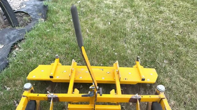 Yellow Lawn Aerator Machine On Grass.Close view of a yellow lawn aerator attachment standing on dry green grass outdoors. Landscaping and turf maintenance equipment detail.