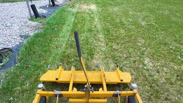 Yellow Lawn Aerator Machine On Grass.Close view of a yellow lawn aerator attachment standing on dry green grass outdoors. Landscaping and turf maintenance equipment detail.