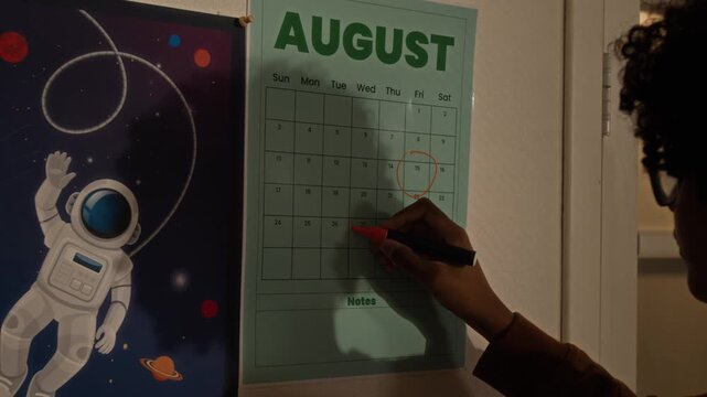 Close-up of motivated young black boy student circling back to school day on August calendar, hanging on wall in bedroom next to space astronaut poster, anticipating return to studies