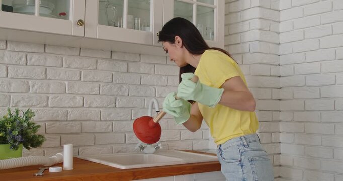 Woman using plunger to unblock kitchen sink clogged at home. Girl homeowner wearing gloves to repair water plumbing and clean, unclog drain blockage before emergency phone call to plumber service