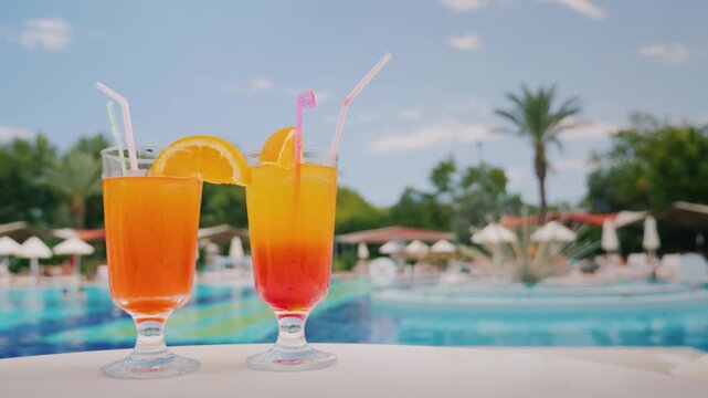 Two vibrant cocktails, one orange, one layered red and yellow, with orange slices and straws, sit on table by sparkling pool with lush palm trees and resort in background