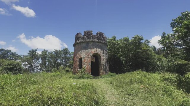 An old round stone tower stands in a grassy clearing surrounded by green trees and bright daylight. The structure has a dark doorway, narrow window openings, reddish stone details, and a weathered cas
