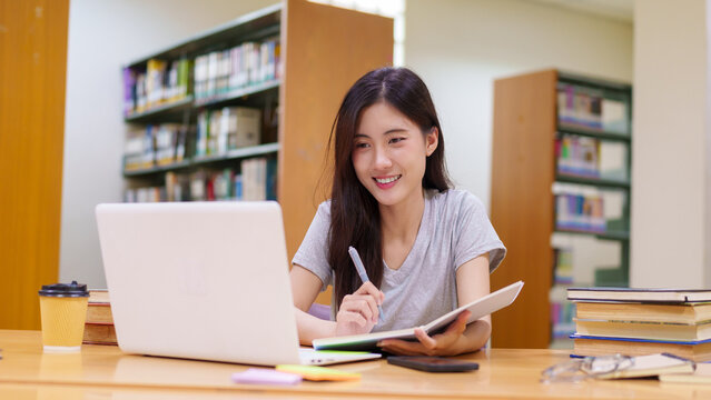 Asian female entrepreneur business student using laptop computer writing notes planning strategy working studying researching at library with books coffee