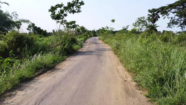 Perspective view of an old rural asphalt road with dirt patches and wild green grass. Scenic look of a lonely rural road surrounded by wild bushes and natural growth. Indian road Potholes. 