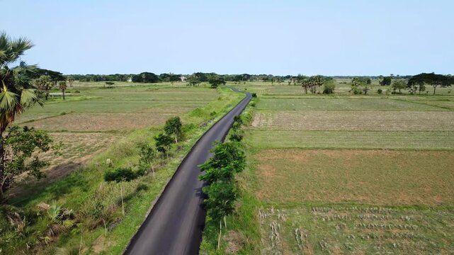 Aerial drone tracking shot of a newly paved rural Indian road winding through green fields. smooth asphalt Indian village road cutting through peaceful countryside.