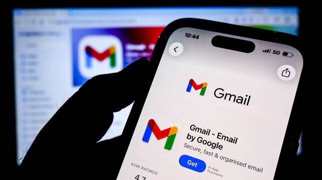 Closeup of a hand holding a smartphone displaying the gmail app download page on the apple app store, with a blurred computer screen in the background