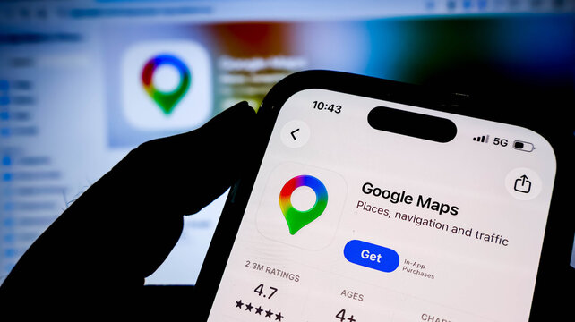Closeup of a hand holding a smartphone displaying the google maps app page on the apple app store, with a blurred background