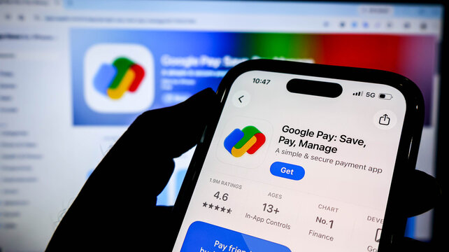 Closeup of a hand holding a smartphone displaying the google pay app on the apple app store, with a blurred computer screen in the background