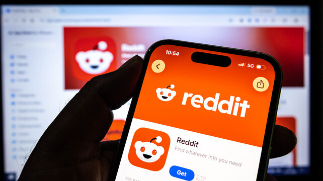 Closeup of a hand holding a smartphone displaying the reddit app store page with a computer monitor in the background showing the same app