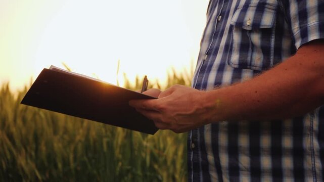 Farmer taking notes on clipboard in wheat field at sunset passing document to another person and shaking hands.