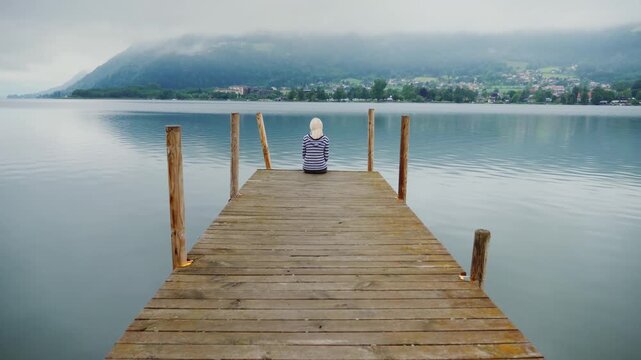 Young person sits on a wooden dock looking out at a vast lake with mountains in the background.