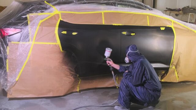 Auto body worker painting masked SUV side panels with spray gun in repair workshop, restoring damaged car surface during professional collision refinishing process