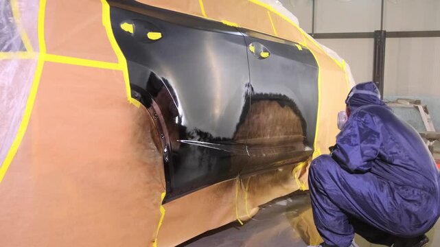 Auto body painter applying black paint with spray gun on masked vehicle panel in repair workshop wide shot, restoring damaged side surface after collision and insurance service