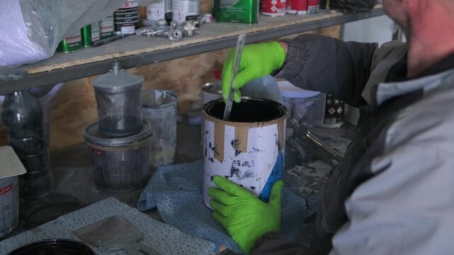 Auto body worker stirring black paint in a mixing can at workshop shelf, preparing coating material for car repainting, collision repair, insurance service and professional refinishing process.