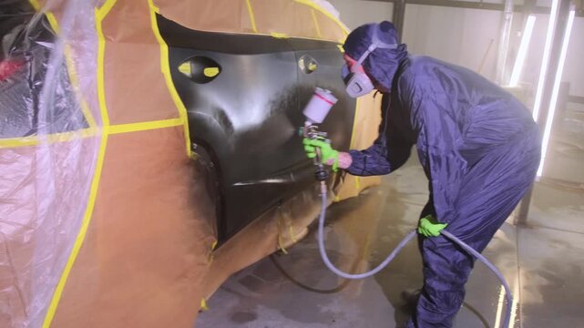 Auto body painter spraying black coating on masked car side panel with paint gun in repair workshop, restoring damaged vehicle surface after accident and insurance repair process