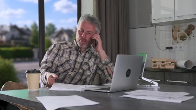 Engineer talking on phone while reviewing documents, working remotely with laptop and construction papers during home office planning and consulting tasks. remote engineer communication