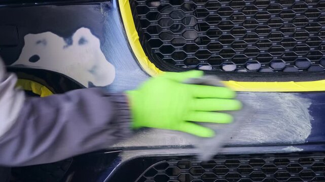 Mechanic spreading repair material across scratched front bumper in automotive bodyshop close-up, restoring damaged coating before paint application and post-accident service inspection