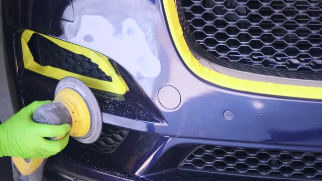 Worker polishing repaired bumper surface with electric buffer in auto paint workshop close-up, preparing front panel for refinishing after collision damage, paint preparation, bumper refinishing