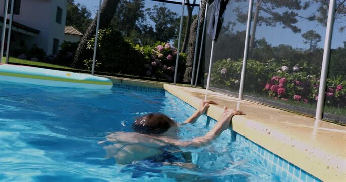 Boy attempting to lift himself out of swimming pool but slipping back in, arms gripping edge with water splashing, near safety fence on sunny backyard day