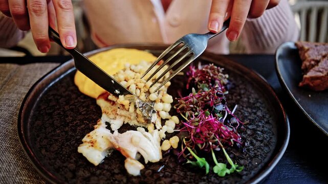 Close up of hands using a fork and knife to cut into a gourmet seared fish fillet with crispy skin, plated with golden puree and microgreens