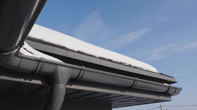 Closeup of dark grey gutter system installed on house roof edge with light snow against clear blue sky.