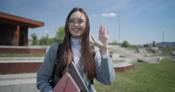 A hand-held portrait of a young female student in casual clothes, wearing glasses and a backpack, holding a notebook and laptop in her hands and making a welcoming gesture with her hands - victory
