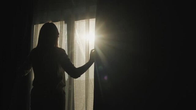 Woman opens curtains in dark bedroom, revealing bright morning sunlight filtering through sheer fabric, creating dramatic silhouette and lens flare.
