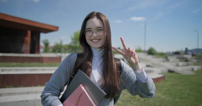 A hand-held portrait of a young female student in casual clothes, wearing glasses and a backpack, holding a notebook and laptop in her hands and making a welcoming gesture with her hands - victory