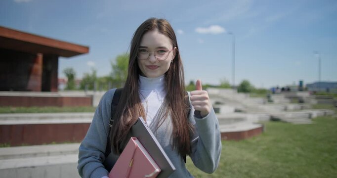 A hand-held portrait of a young female student in casual clothes, wearing glasses and a backpack, holding a notebook and laptop in her hands and making a welcoming gesture with her hands - victory