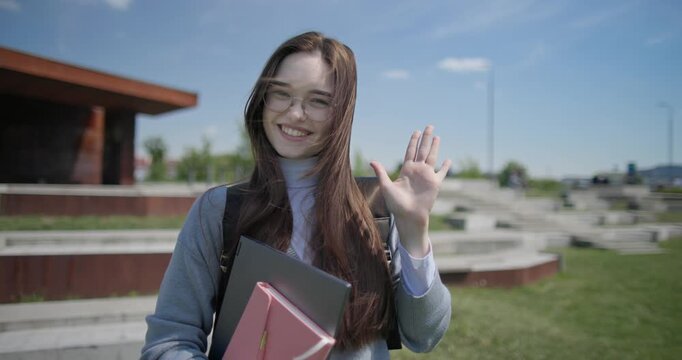 A hand-held portrait of a young female student in casual clothes, wearing glasses and a backpack, holding a notebook and laptop in her hands and making a welcoming gesture with her hands - victory