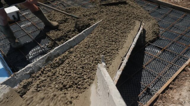 Fresh concrete flows down metal chute onto rebar reinforced construction site, worker spreads with rake.