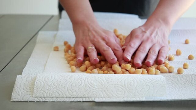 Close-up of a woman's hands using kitchen paper to dry cooked chickpeas thoroughly on a flat surface