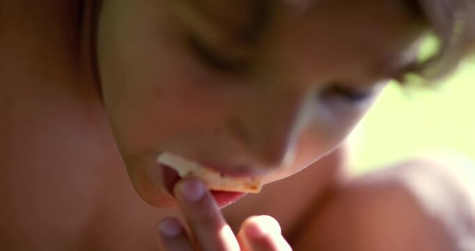 Closeup of shirtless boy eating piece of bread outdoors, hand raised to mouth as he takes a bite during relaxed summer moment in grassy yard