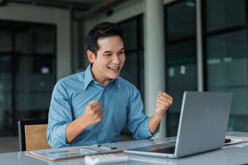 Fototapeta premium Happy business man cheering, achieving goals, and feeling victorious while working on a laptop in a modern office