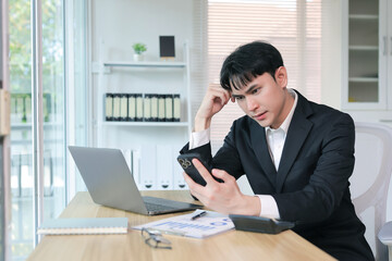 Naklejka premium Young Asian businessman sitting at office desk, looking worried and frustrated while holding a phone and processing information